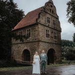 Brautpaar steht vor einer historischen Burg und lächelt händchenhaltend in die Kamera