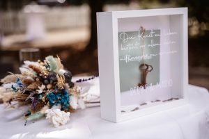 Auf einem weißen Beistelltisch am Trauort befindet sich das vorberreietet Trauritual "Schlüssel zum Herzen" neben dem Brautstrauß